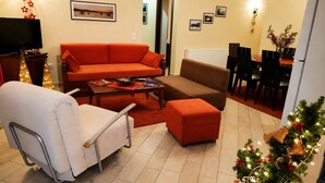 A living room with a sofa, ottoman, and a white chair. There is a dining area with chairs and a table. A television is mounted on the wall. - Zidoron House - Peaceful Retreat at Lake Kerkini (Mandraki)