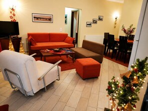 Una sala de estar con un sofá, un reposapiés y una silla blanca. Hay un comedor con sillas y una mesa. Un televisor está montado en la pared.