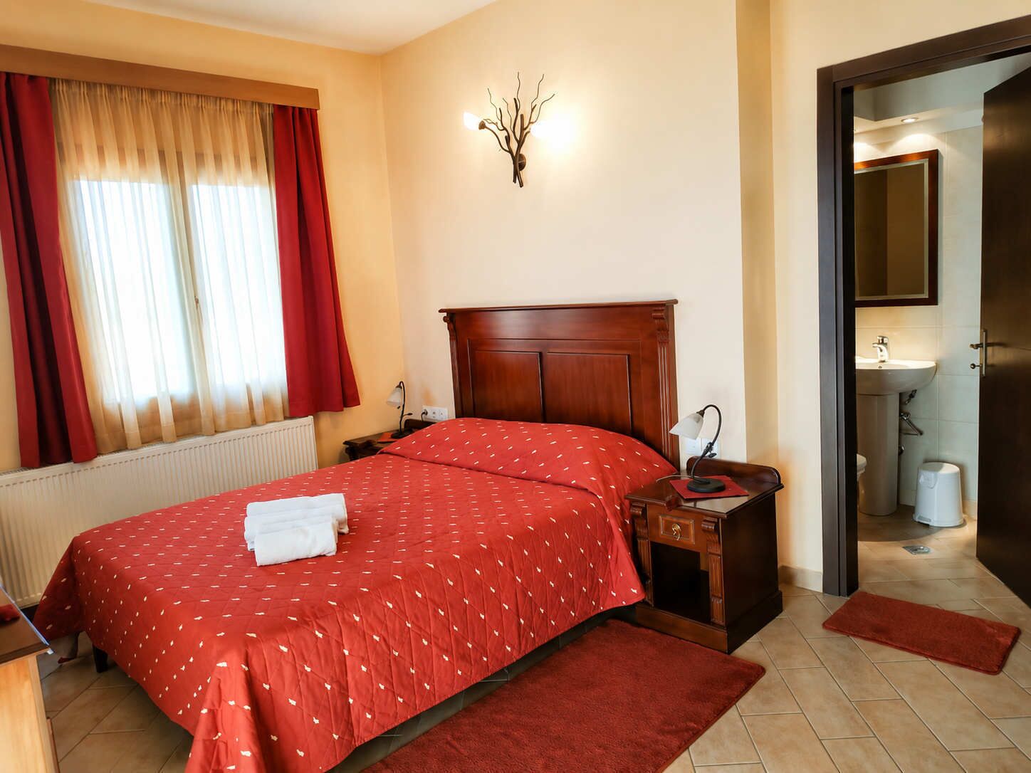 A hotel room with a bed, a nightstand, a wall-mounted lamp, a radiator, a window with curtains, and a door leading to a bathroom.