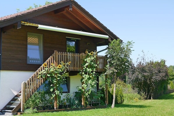 Una casa de madera de dos pisos, con balcón, una escalera visible y un jardín con girasoles.