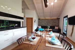 Una cocina moderna con armarios blancos, una mesa de comedor para cuatro personas con posavasos y una sala de estar con sofá y televisión.