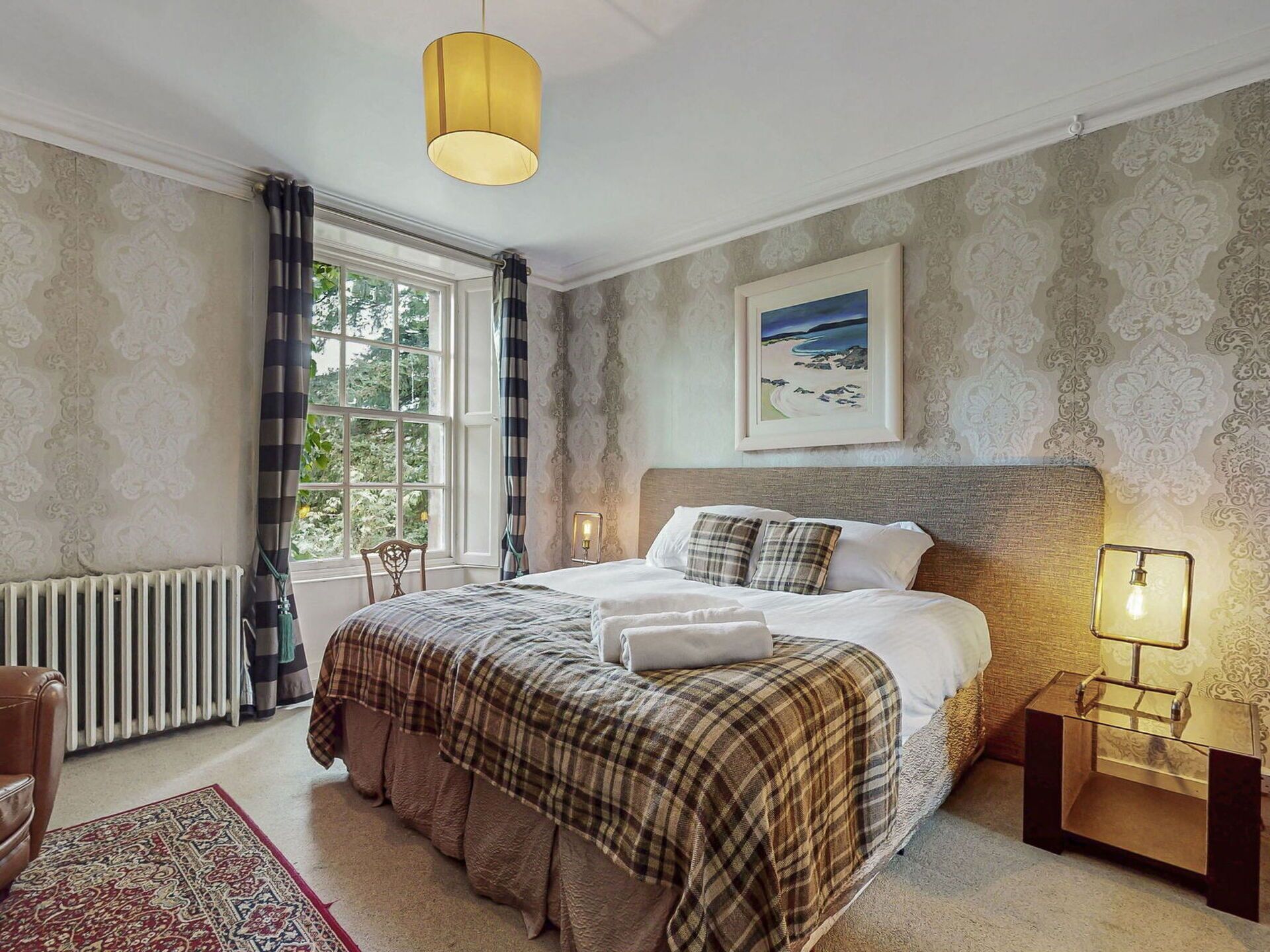 Une chambre avec un grand lit, une table de chevet avec une lampe, un radiateur, une fenêtre avec des rideaux et un tableau encadré au mur.