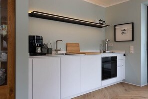 A modern kitchen with white cabinetry, a built-in oven, a coffee machine, and a wooden cutting board. - Holiday apartment in Wildeshausen with Wi-Fi (Wildeshausen)