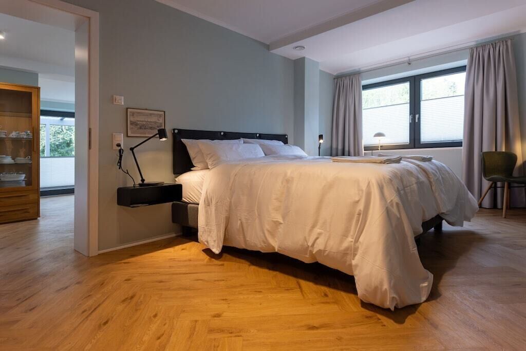 Ein Schlafzimmer mit einem großen Bett, einem Nachttisch, einer Lampe und einem Fenster mit Vorhängen.