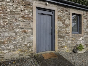 Ein Steinhäuschen mit einer grauen Tür und einem Fenster. Ein Schild über der Tür trägt die Aufschrift 'Garden Cottage'.