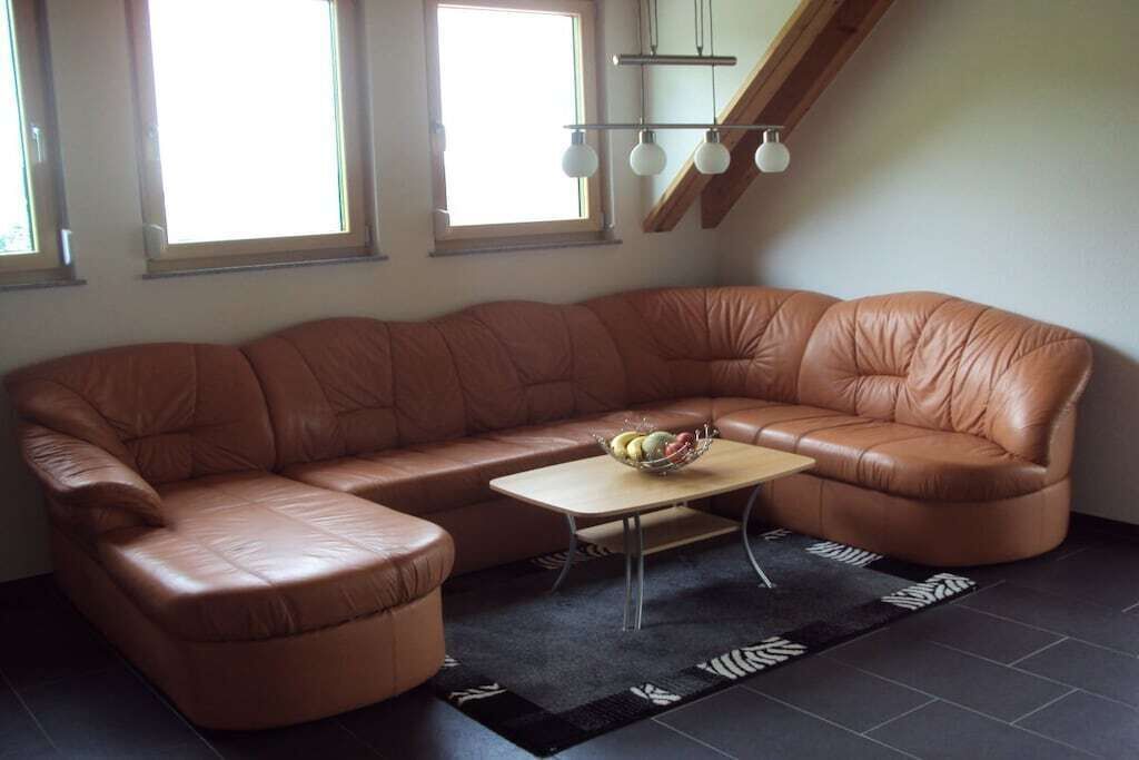 Ein braunes, ledernes Ecksofa mit einem kleinen Tisch und einer Obstschale, ein schwarzer Teppich mit Blattmuster und eine Treppe mit Holzbalken und Pendelleuchten.