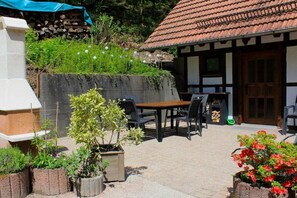 An outdoor patio with a table and chairs, surrounded by potted plants and a building with a tiled roof. - ||------------------storage building------------|| (Hornberg)