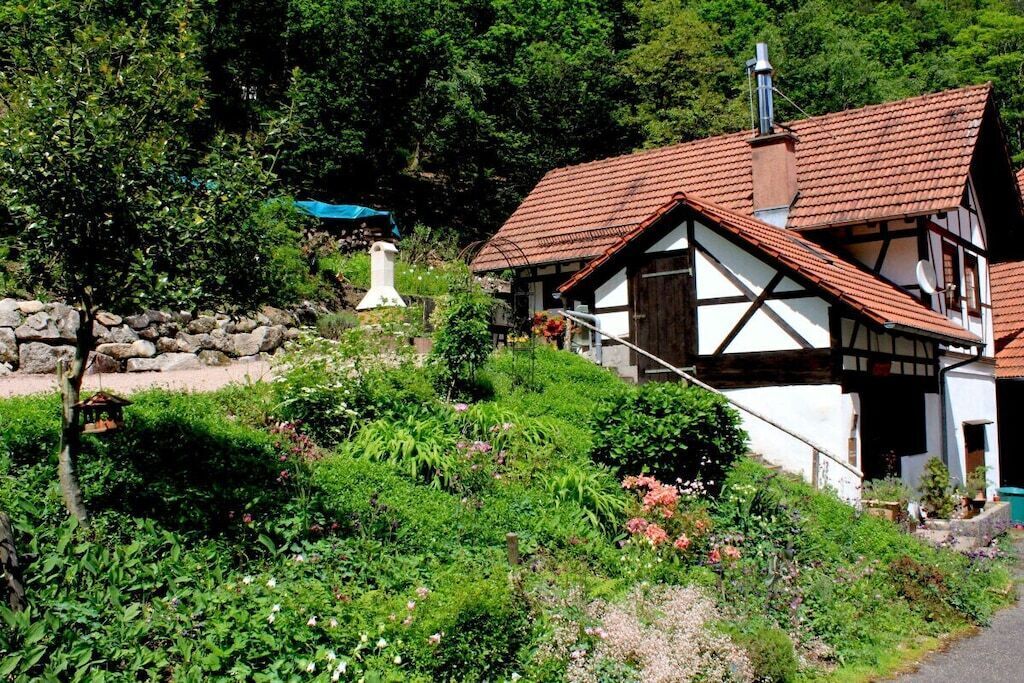 A traditional half-timbered house with a sloped roof, surrounded by lush greenery and a stone wall.