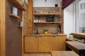 Een kleine keuken met houten keukenkastjes, een spoelbak en een magnetron. Boven het aanrecht staan planken met glazen en een plant.