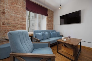 Een moderne woonkamer met een bakstenen muur, een blauwe bank, een houten fauteuil, een salontafel en een aan de muur bevestigde televisie.