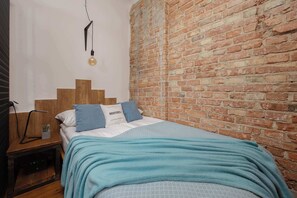 Een slaapkamer met een bakstenen muur, een houten hoofdbord, een bed met een blauwe deken en een nachtkastje met een lamp en een klein plantje.