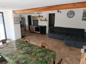Ein Essbereich mit Tisch, der mit einem tropischen Blattmuster bedeckt ist, einer grauen Couch und einer Küche im Hintergrund.