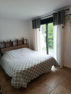 Une chambre à coucher avec un lit, une fenêtre avec des rideaux, une étagère ornée d’objets décoratifs et une vue sur la verdure à l’extérieur.