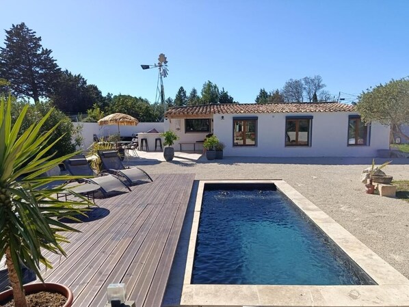 Une piscine avec une terrasse en bois, des chaises longues et un bâtiment blanc doté d’un toit recouvert de tuiles.