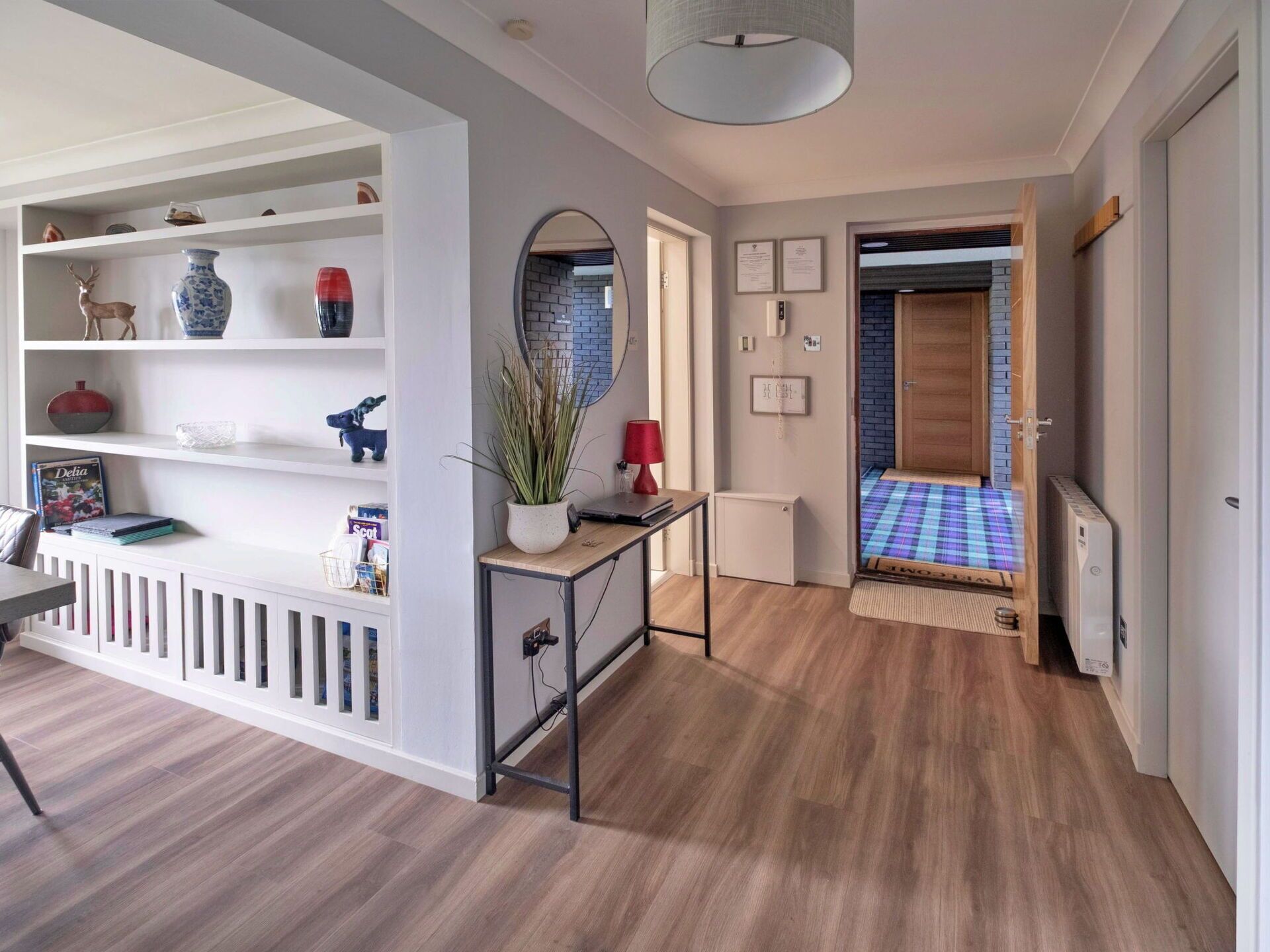 A modern hallway with a white console table, shelves with decorative items, a red lamp, and a door leading to another room.
