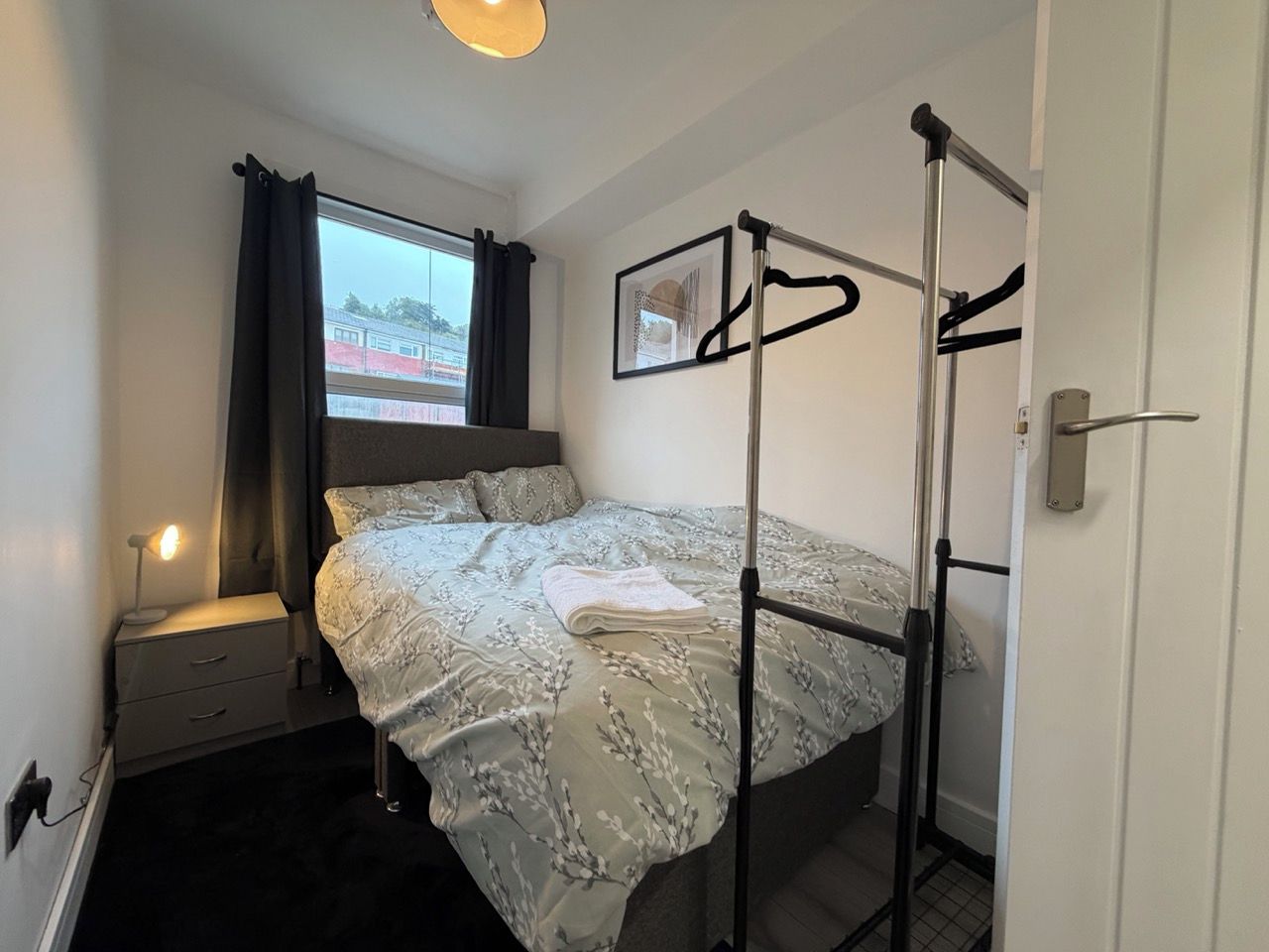 Ein Einzelbett mit Metallgestell, ein Nachttisch mit Lampe, ein Fenster mit Vorhängen und eine Tür mit Griff.