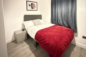 Une chambre à coucher avec un lit, une table de chevet, un tiroir, une couverture rouge, une tête de lit grise, un rideau gris et un tableau encadré accroché au mur.
