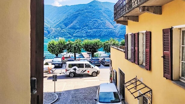 A street view with parked cars, a building with a balcony, and a mountainous landscape in the background. - Lovely apartment for 2 people (Porlezza)