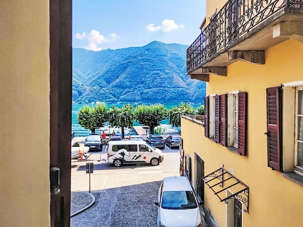 A street view with parked cars, a building with a balcony, and a mountainous landscape in the background. - Beautiful apartment for 2 guests (Porlezza)