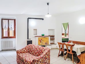 A living room with a sofa, a dining table with chairs, a radiator, a ceiling light fixture, and a window with curtains. - Cosy apartment for 2 guests (Porlezza)