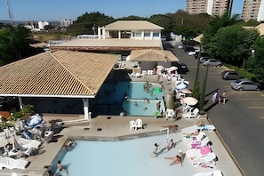 Ein Schwimmbecken mit Liegestühlen und Sonnenschirmen, umgeben von einem Parkplatz und Gebäuden.