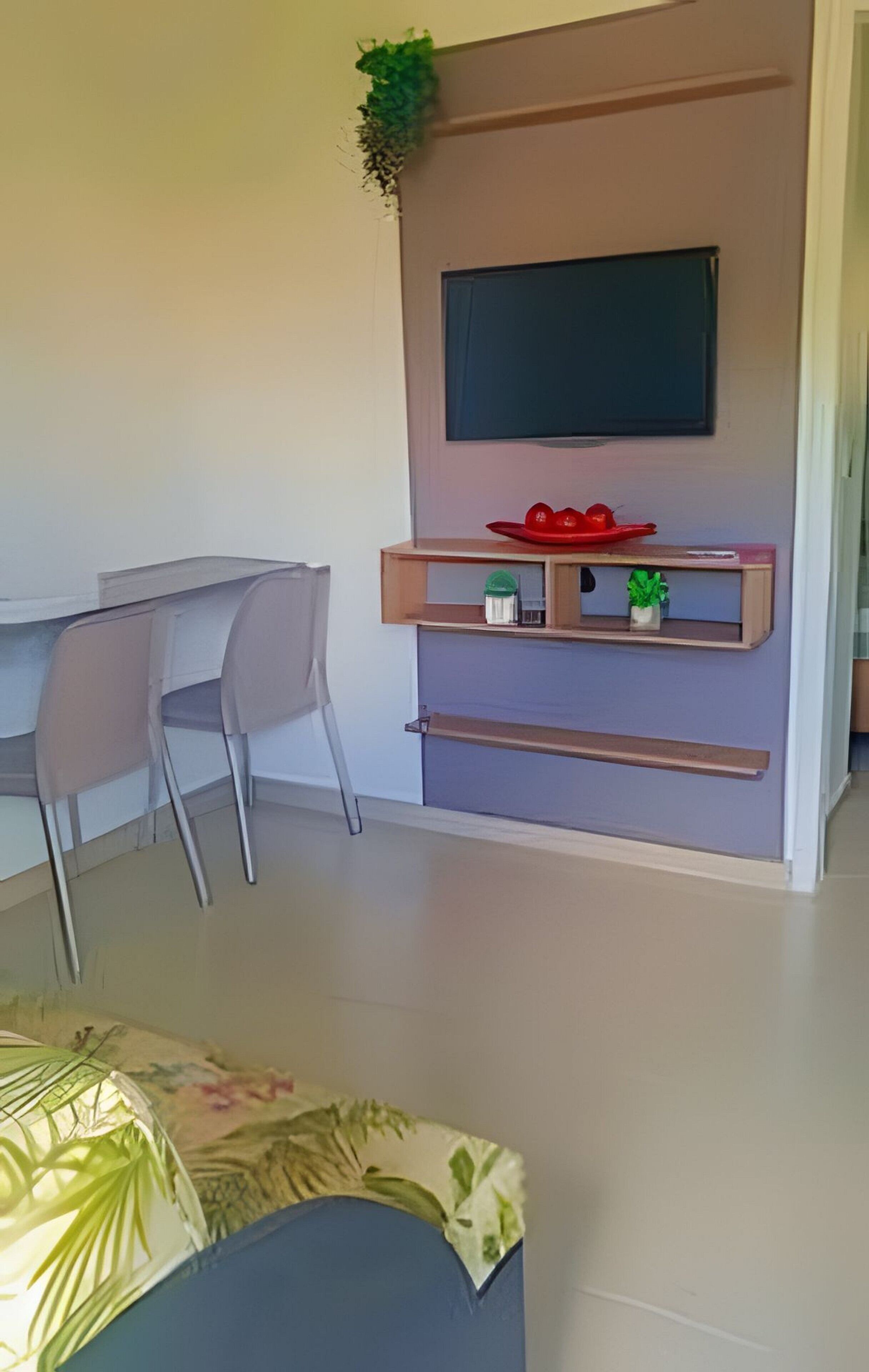 A modern living room with a flat-screen TV mounted on a wall, a dining table with chairs, and a small shelf with decorative items.