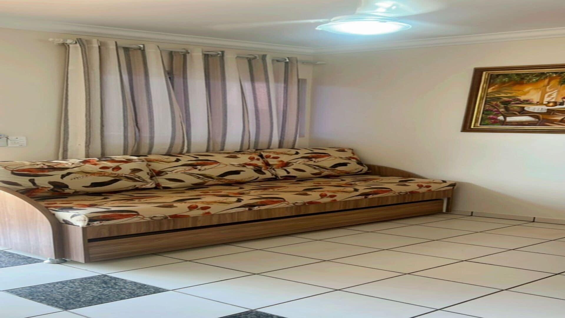 Una habitación con sofá cama, cortinas, un cuadro y suelo de baldosas.