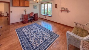 Una habitación amplia con pisos de madera, un tapete grande con diseño azul, una silla de mimbre, un escritorio con silla y una ventana con vistas.