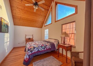 A bedroom with a wooden ceiling, a bed with a quilt, a nightstand, a lamp, and a window with curtains.
