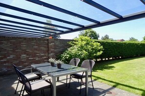 Eine überdachte Terrasse mit Tisch und Stühlen, umgeben von einer Ziegelmauer und einem Hecken.