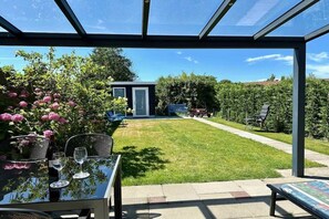 Eine verglaste Terrasse mit einem Tisch für zwei Personen, eine kleine Gartenhütte und ein gepflegter Rasen.