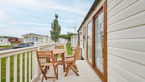 A wooden deck with a table and chairs, a white railing, and a view of other mobile homes in the background.