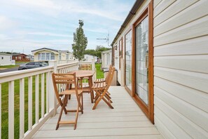 A wooden deck with a table and chairs, a white railing, and a view of other mobile homes in the background.