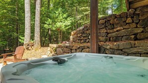 A hot tub in a forested area with a stone wall and a wooden chair. - River Resort | Hot Tub + Game Room + Fenced Yard (Ellijay)
