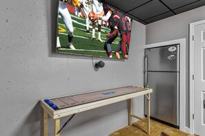 Une pièce avec un téléviseur fixé au mur diffusant un match de football, une table en bois avec un jeu de société, un réfrigérateur et une porte.