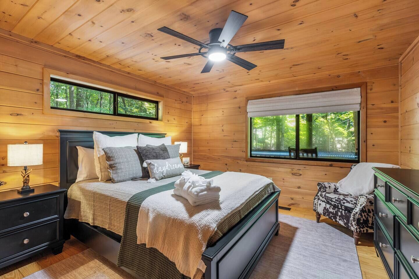 Ein Schlafzimmer einer Holzhütte mit einem Bett, Nachttischen, einer Kommode, einem Sessel und einem Fenster mit Blick auf Bäume.