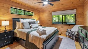 A wooden cabin bedroom with a bed, nightstands, a dresser, a chair, and a window with a view of trees. - River Resort | Hot Tub + Game Room + Fenced Yard (Ellijay)
