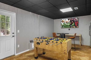 A room with a foosball table, a television mounted on the wall, and a door with a window. - River Resort | Hot Tub + Game Room + Fenced Yard (Ellijay)