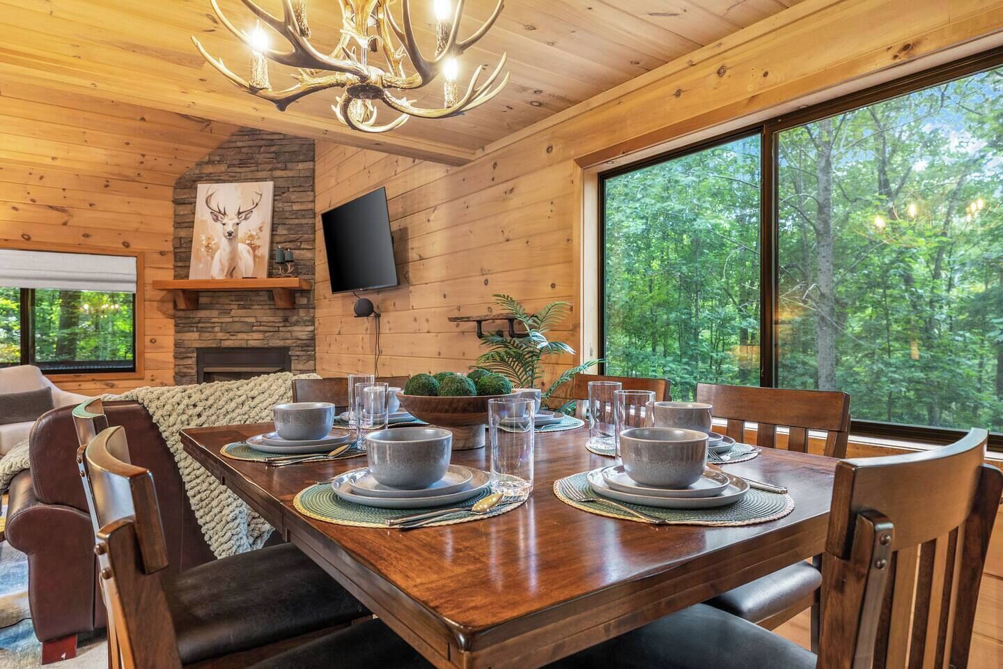 A wooden dining room with a large table set for six, a stone fireplace, and a television mounted on the wall.