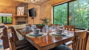 A wooden dining room with a large table set for six, a stone fireplace, and a television mounted on the wall. - River Resort | Hot Tub + Game Room + Fenced Yard (Ellijay)