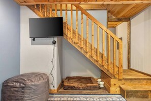 A room with a wooden staircase, a mounted television, a cushioned seating area, and a patterned rug. - River Resort | Hot Tub + Game Room + Fenced Yard (Ellijay)