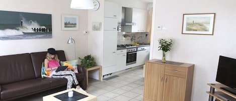 Una sala de estar con sofá, una mesa de centro y televisión. Hay una zona de cocina con armarios y estufa.