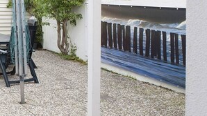 A patio area with a metal table and chairs, a tree, and a wall with a mural of a wooden fence and waves. - Holiday home in Domburg with swimming pool (domburg)
