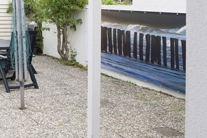 Een terras met een metalen tafel en stoelen, een boom en een muur met een muurschildering van een houten omheining en golven.