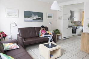 Een woonkamer met een bruine bank, een klein houten bijzettafeltje en een schilderij van een zeegezicht aan de muur.