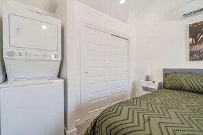 A hotel room with a washing machine, a bed with a patterned bedspread, a nightstand with a lamp, and a door.