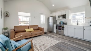 Uma sala de estar ampla com um sofá de couro marrom, uma poltrona azul e uma mesa de centro de madeira. Uma cozinha com armários brancos e eletrodomésticos de aço inoxidável pode ser vista ao fundo.