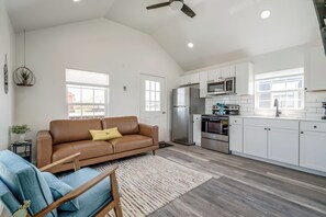 A spacious living room with a brown leather sofa, a blue armchair, and a wooden coffee table. A kitchen with white cabinets and stainless steel appliances is visible in the background. - Cottage on Historic Rt. 66 Moonstone (El Reno)