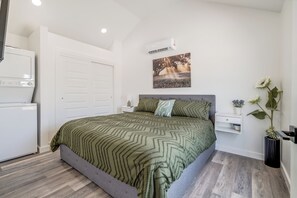 A bedroom with a bed, a nightstand, a wall-mounted air conditioner, and a door. - Cottage on Historic Rt. 66 Moonstone (El Reno)