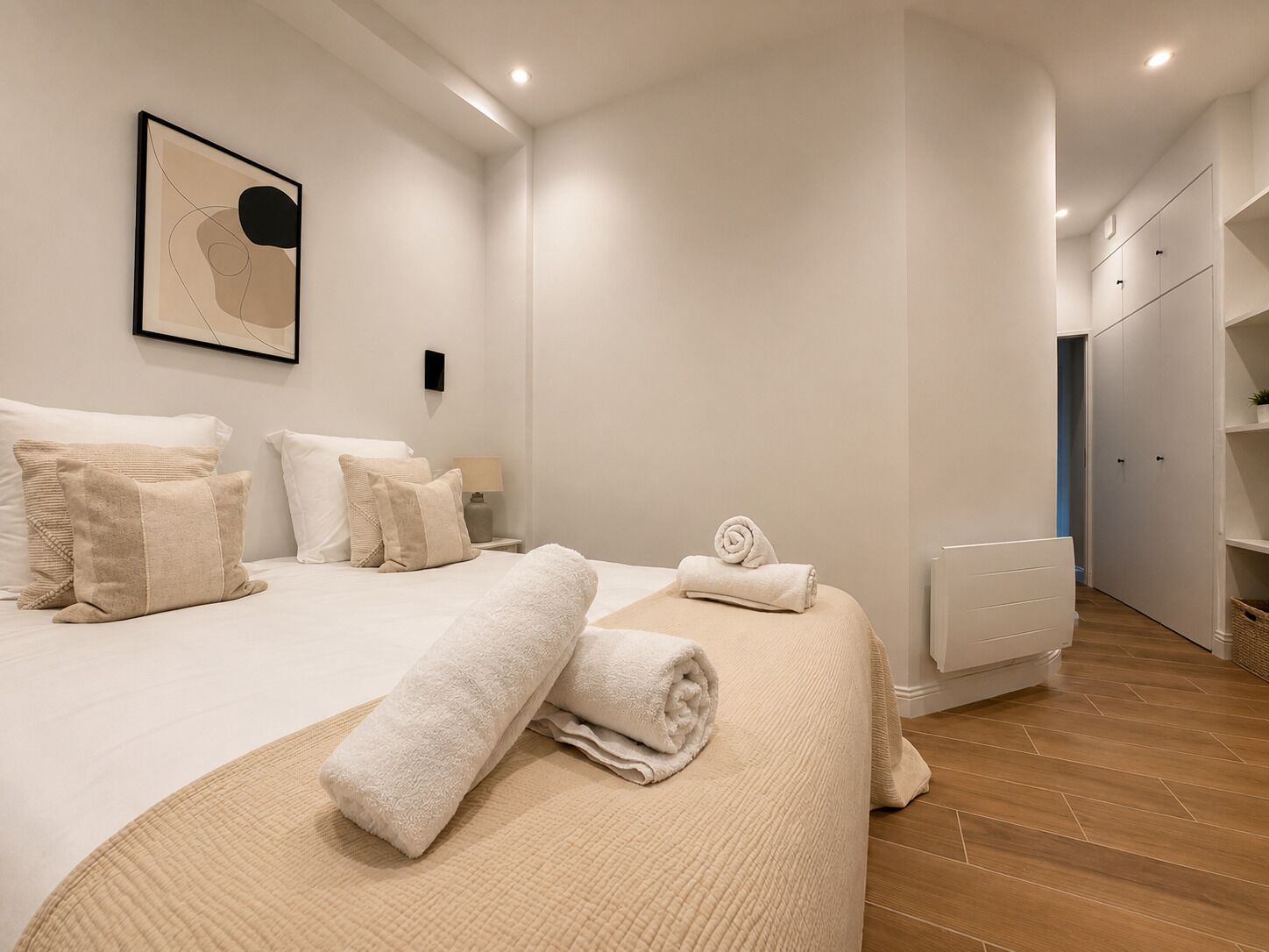 Ein modernes Schlafzimmer mit einem Bett, Kissen, einem zusammengerollten Handtuch, einem Nachttisch mit Lampe und einem Wandbild.